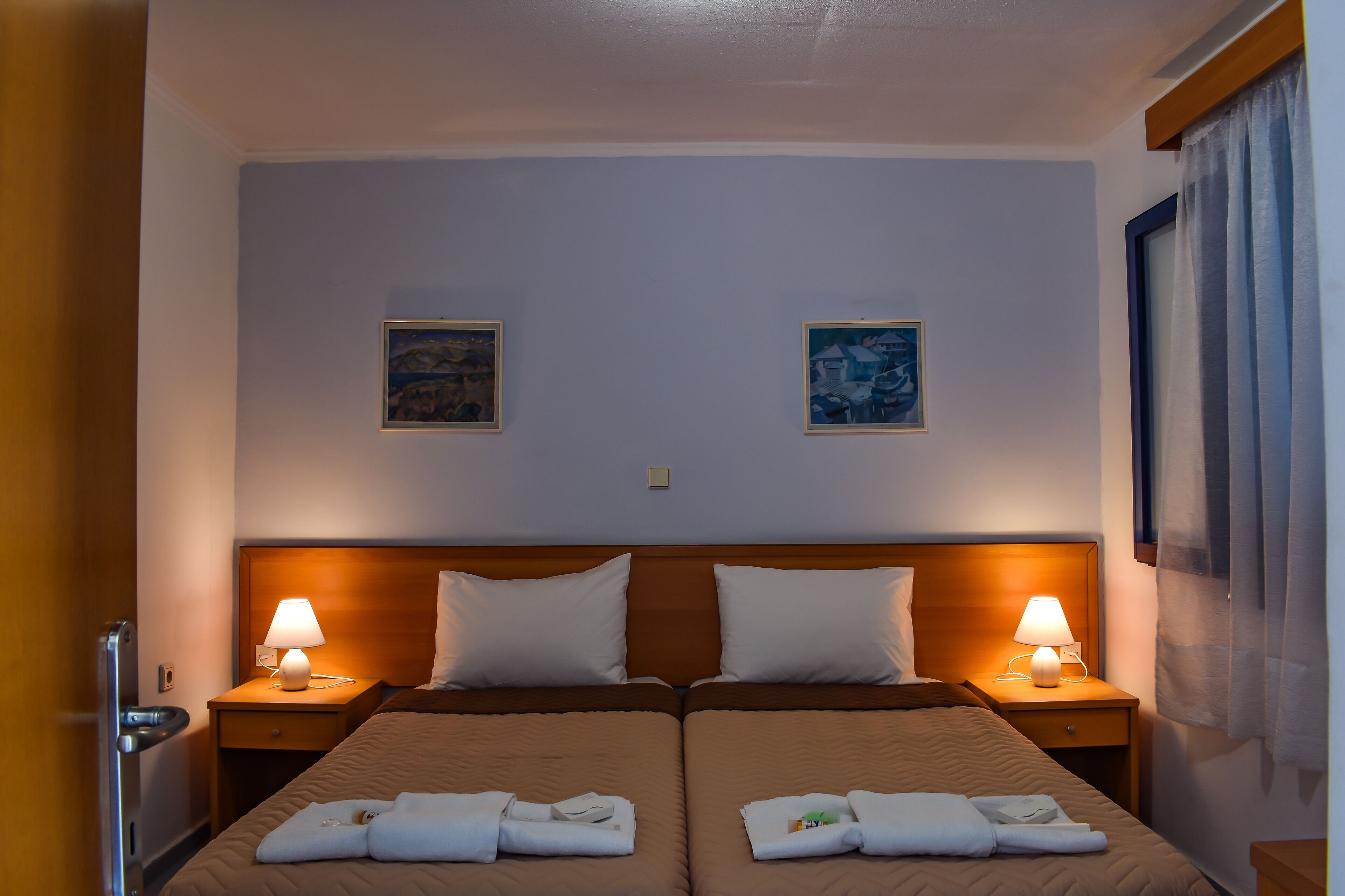 Een hotelkamer met twee bedden, elk voorzien van een kussen en een opgevouwen handdoek. Twee nachtlampjes en twee ingekaderde foto’s hangen aan de muur.