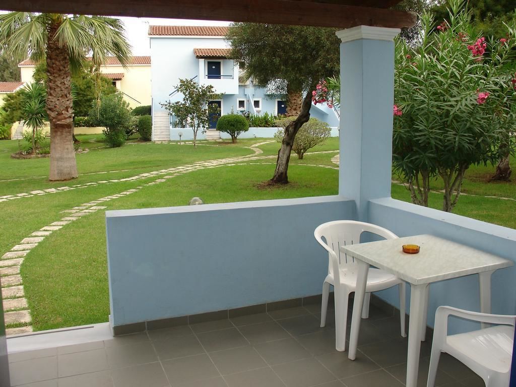 A balcony with a table and two chairs, overlooking a well-maintained garden with a pathway and a building in the background.