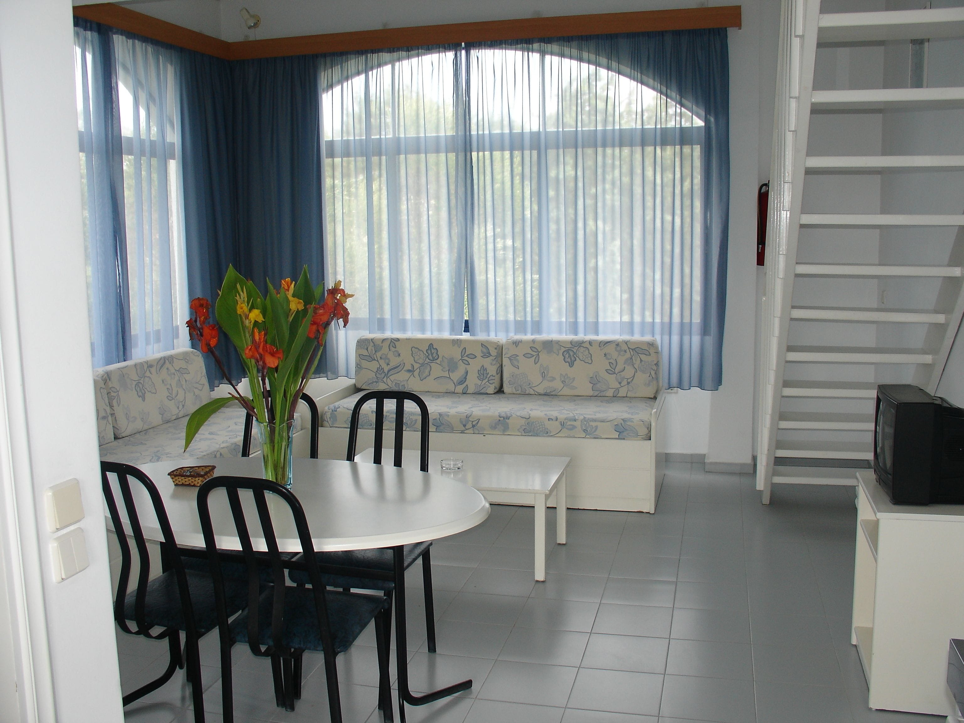A dining area with a table, chairs, a vase with flowers, a sofa with floral upholstery, and a staircase.