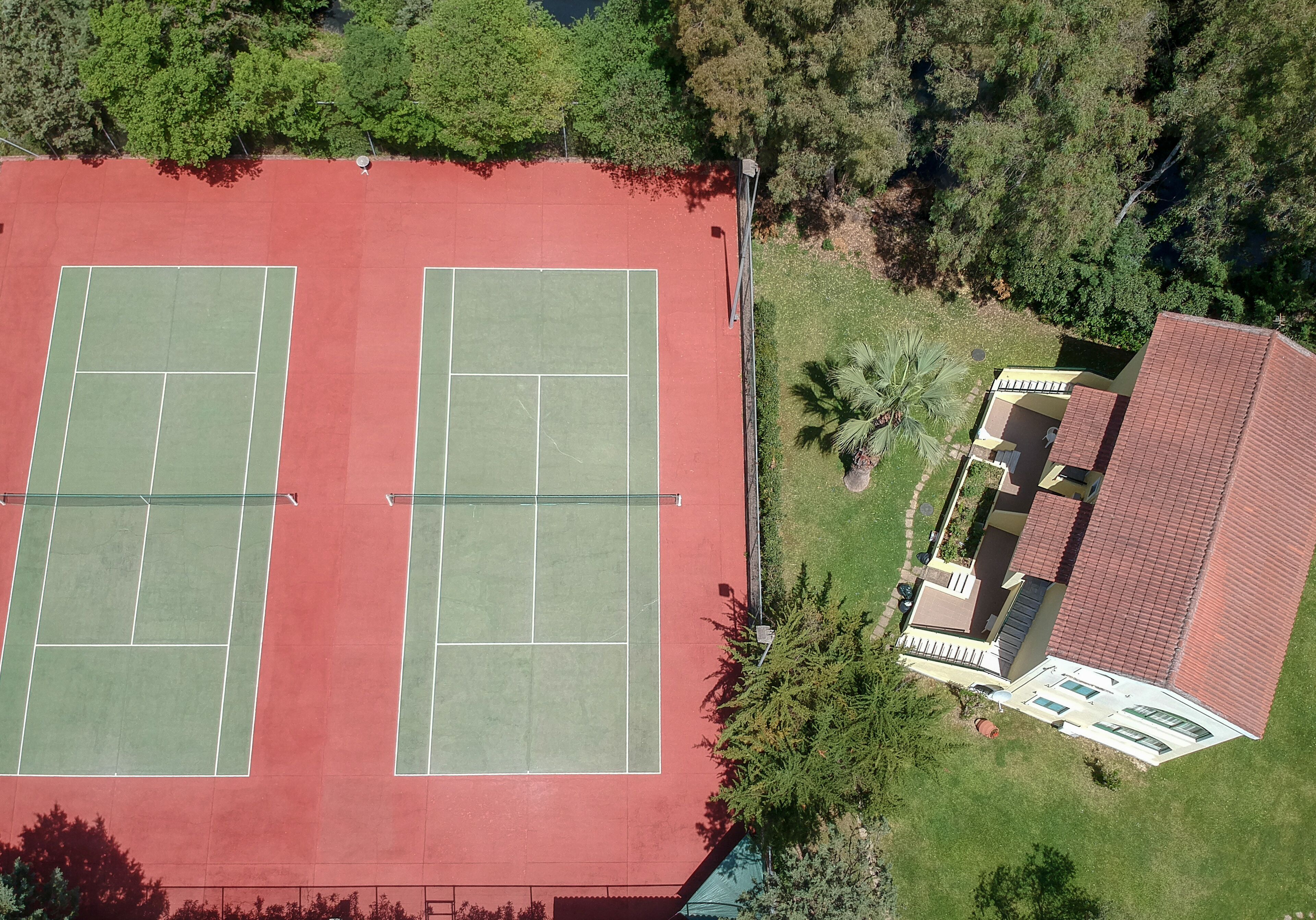Vista aérea de una cancha de tenis con dos canchas, rodeadas de árboles y un edificio con tejado rojo.