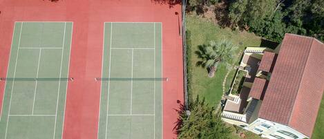 Vista aérea de una cancha de tenis con dos canchas, rodeadas de árboles y un edificio con tejado rojo.