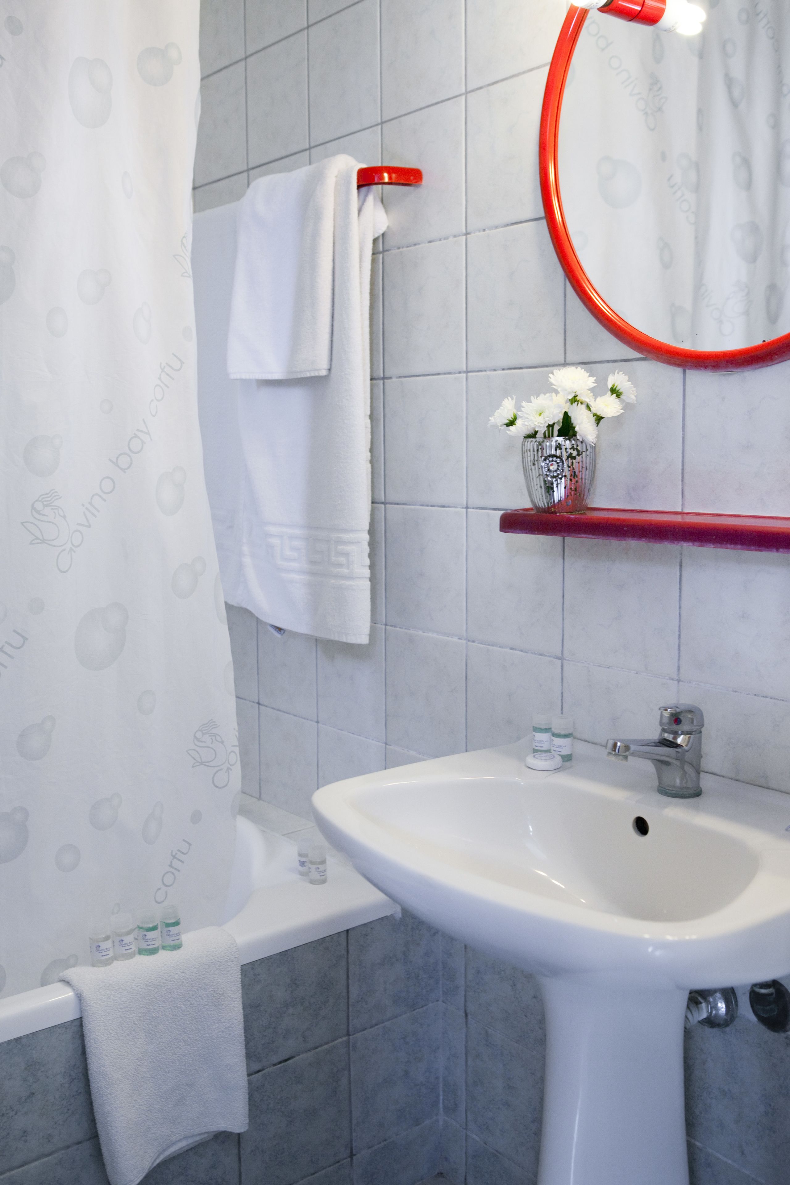 Un bagno con un lavandino bianco, uno specchio con cornice rossa, un asciugamano bianco appeso al muro e una tenda da doccia decorata.