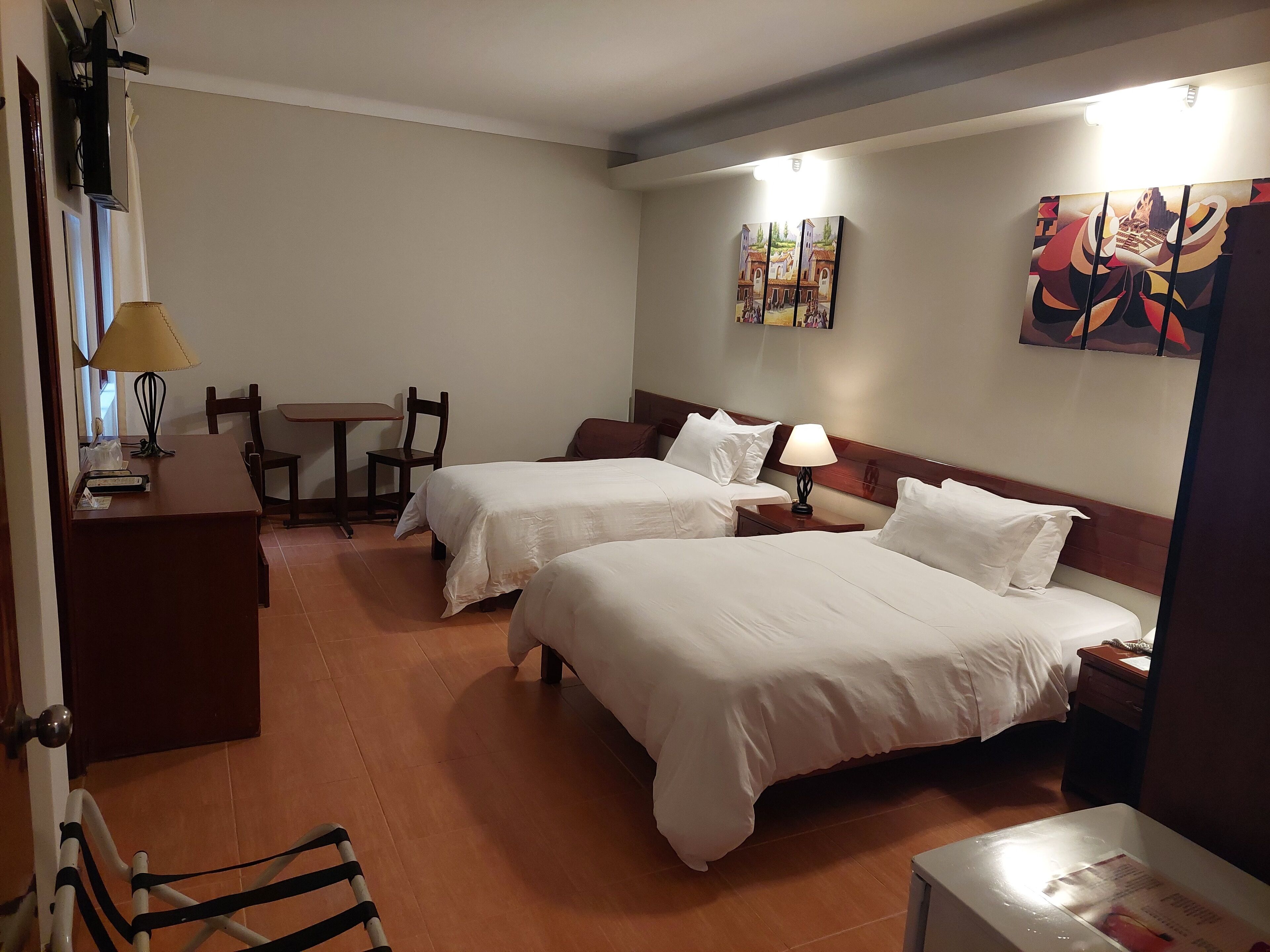 A hotel room with two beds, a desk, a chair, a lamp, and artwork on the walls.