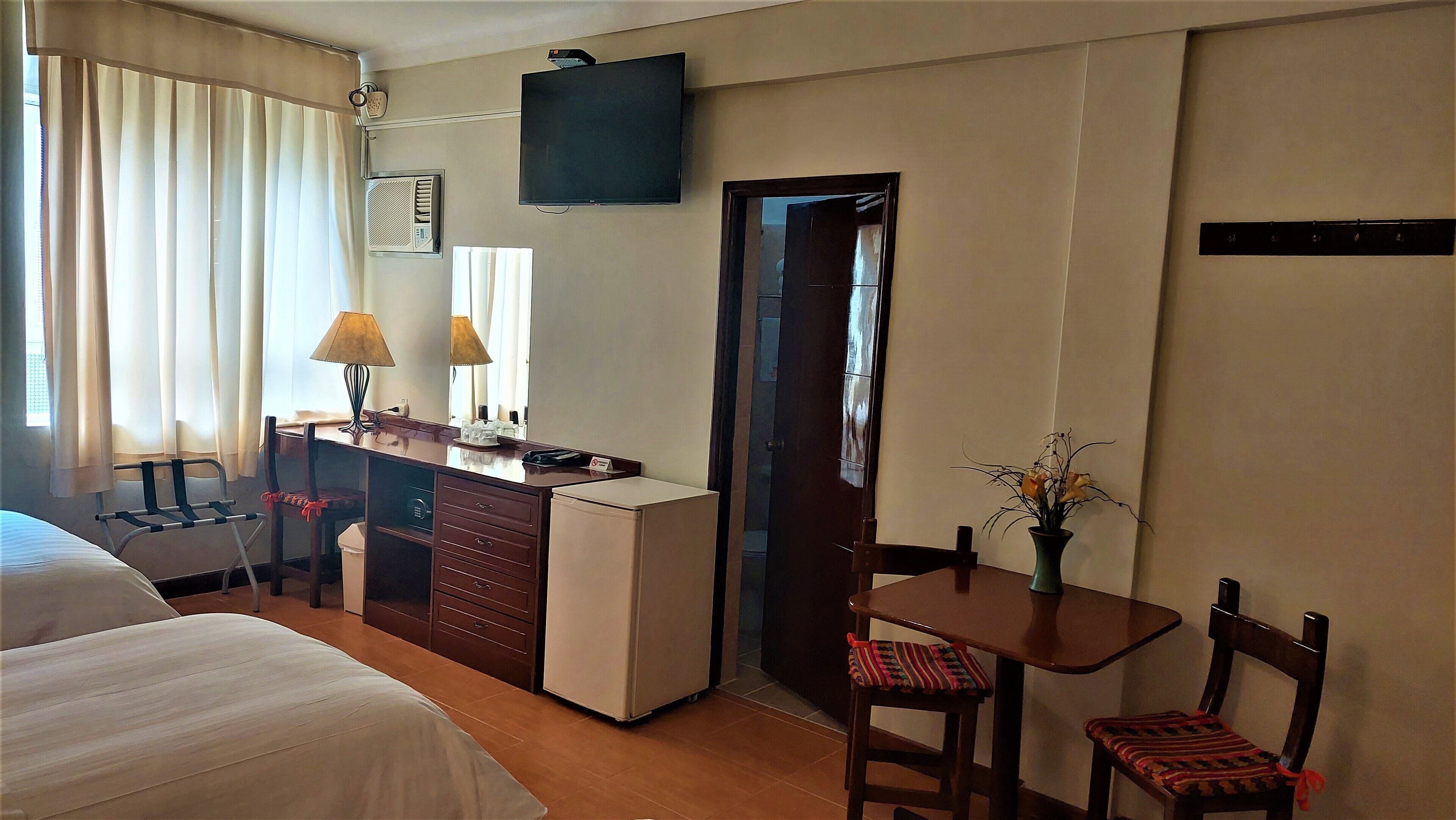 A hotel room with a bed, a desk with a lamp, a television, a small table with a vase, and a chair.