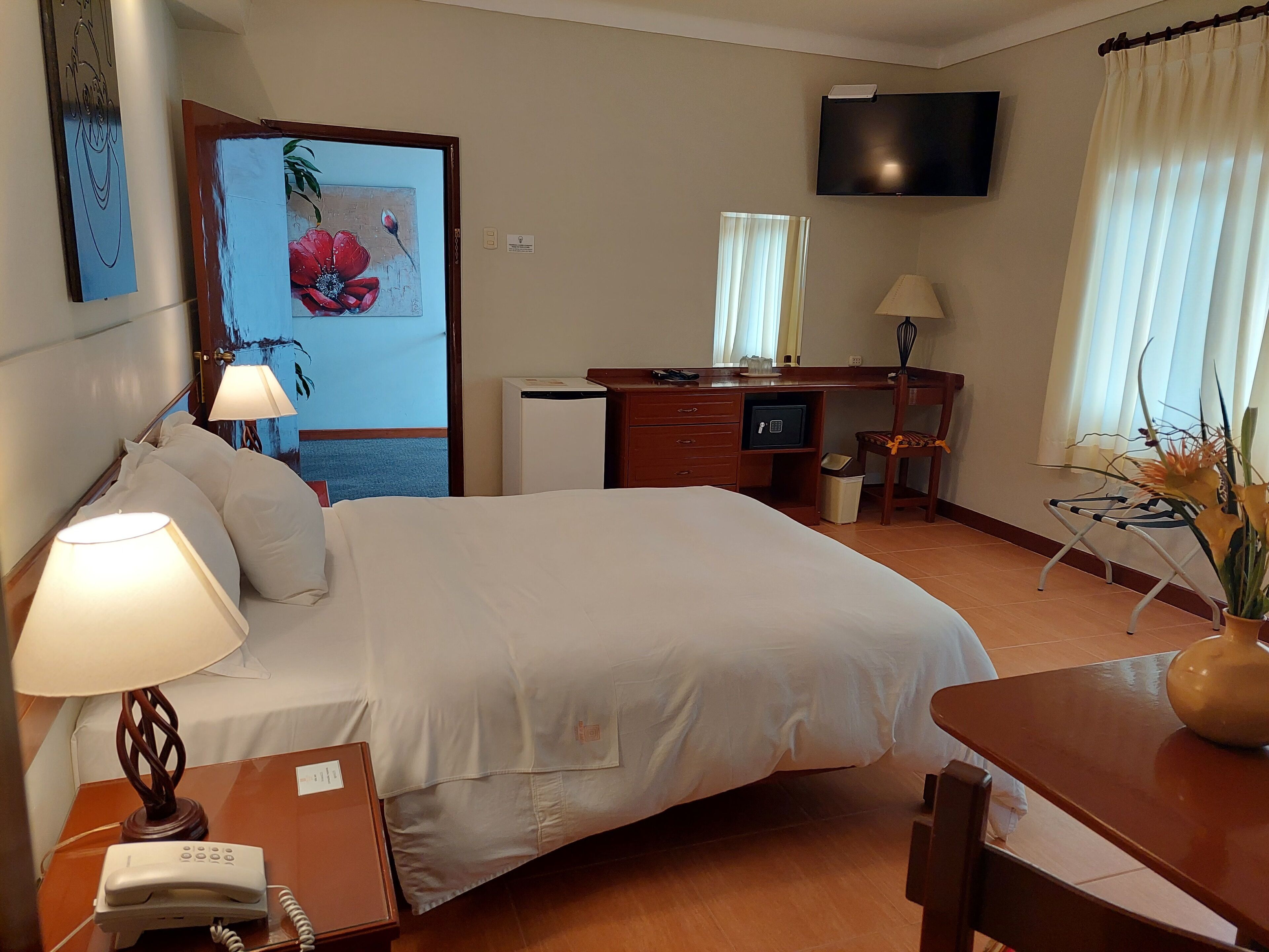A hotel room with a bed, desk, chair, lamp, phone, TV, and a painting on the wall.