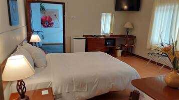 A hotel room with a bed, desk, chair, lamp, phone, TV, and a painting on the wall.