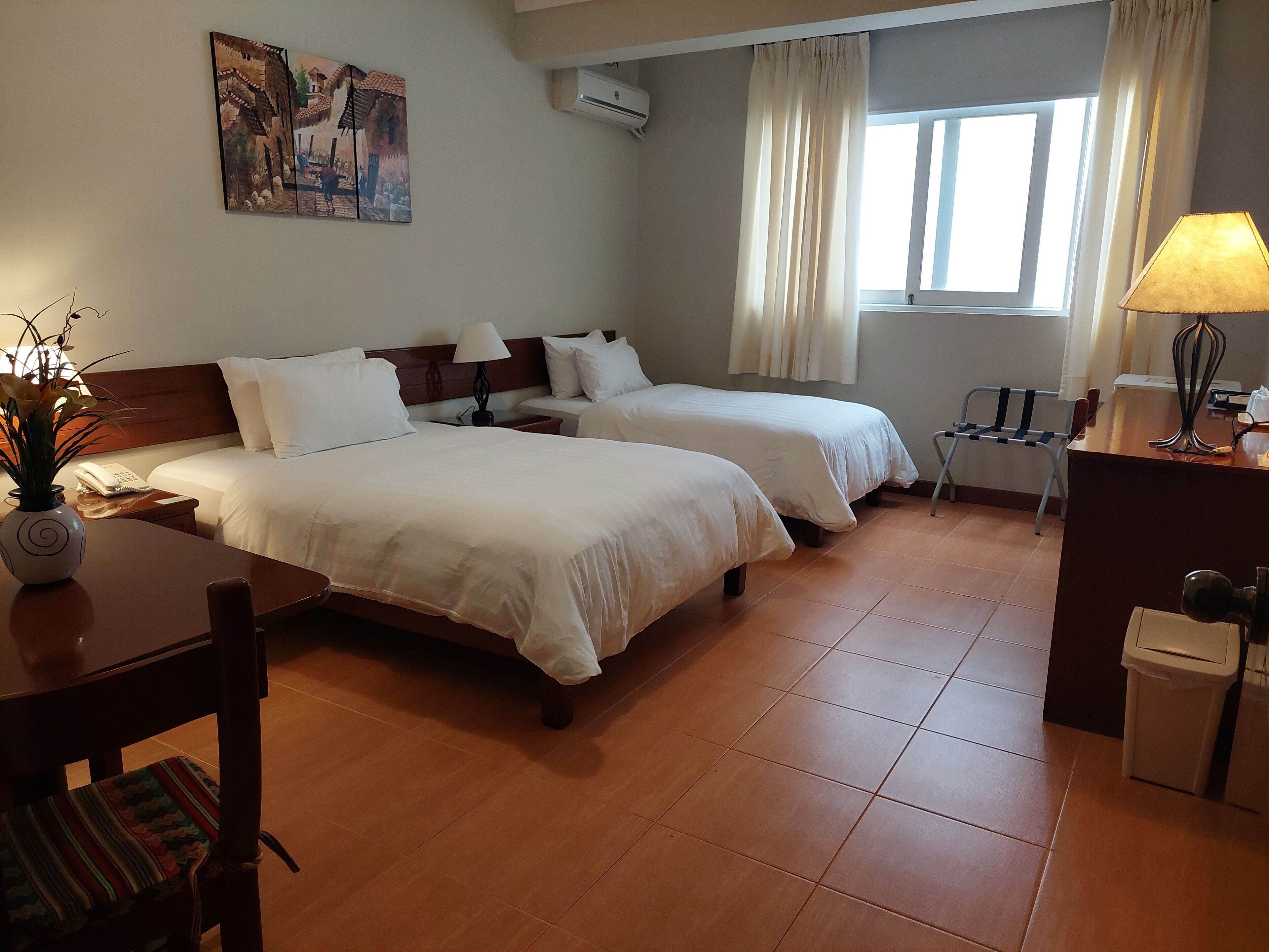 A hotel room with two beds, a desk with a lamp, a chair, a small table with a vase, and a painting on the wall.