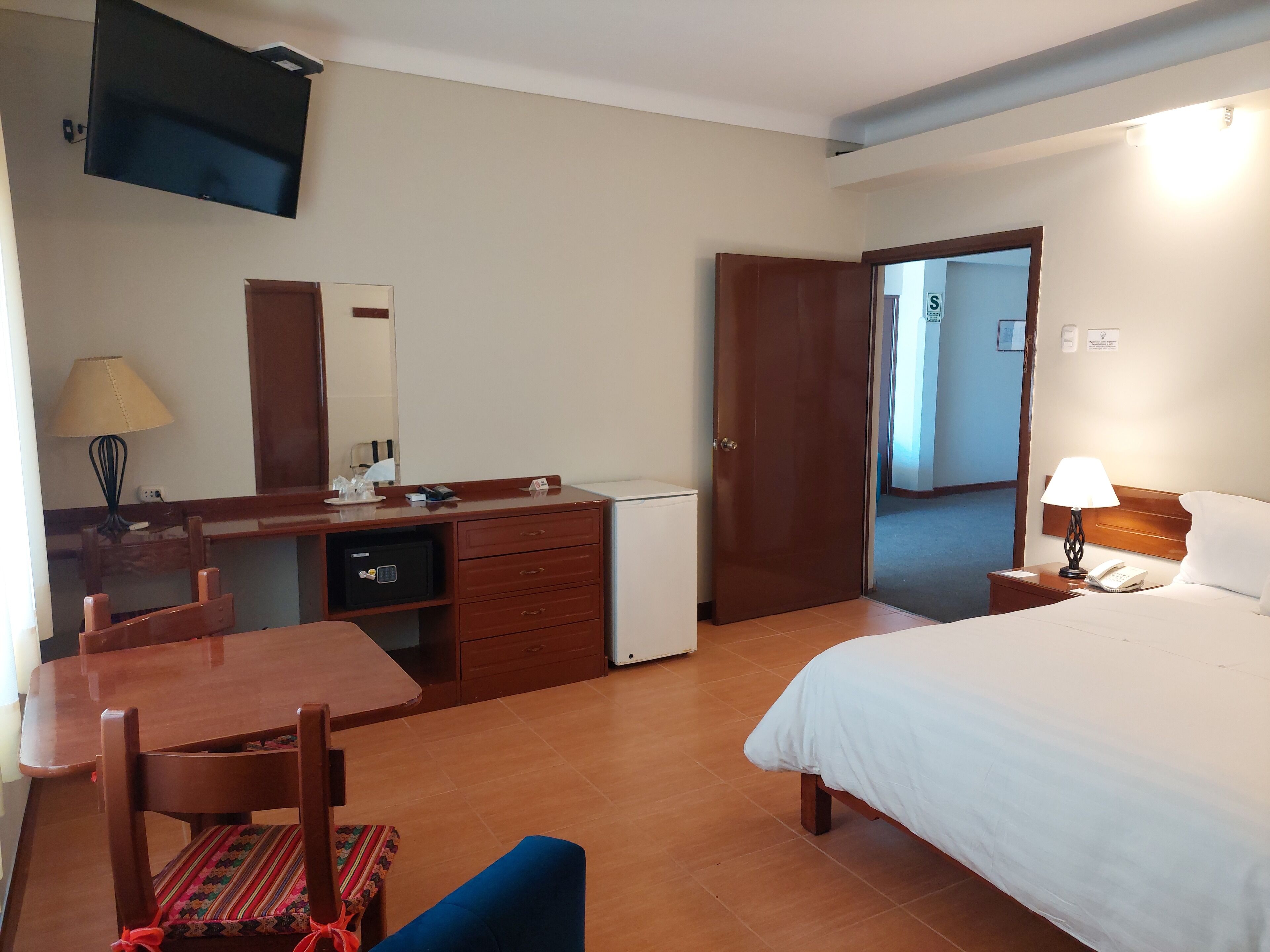 A hotel room with a bed, a desk with a chair, a television, a lamp, and a door leading to another room.