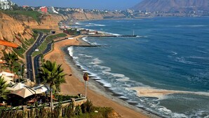 Un hotel frente al mar con vistas a la playa, una carretera serpenteando por la costa y un faro a lo lejos.