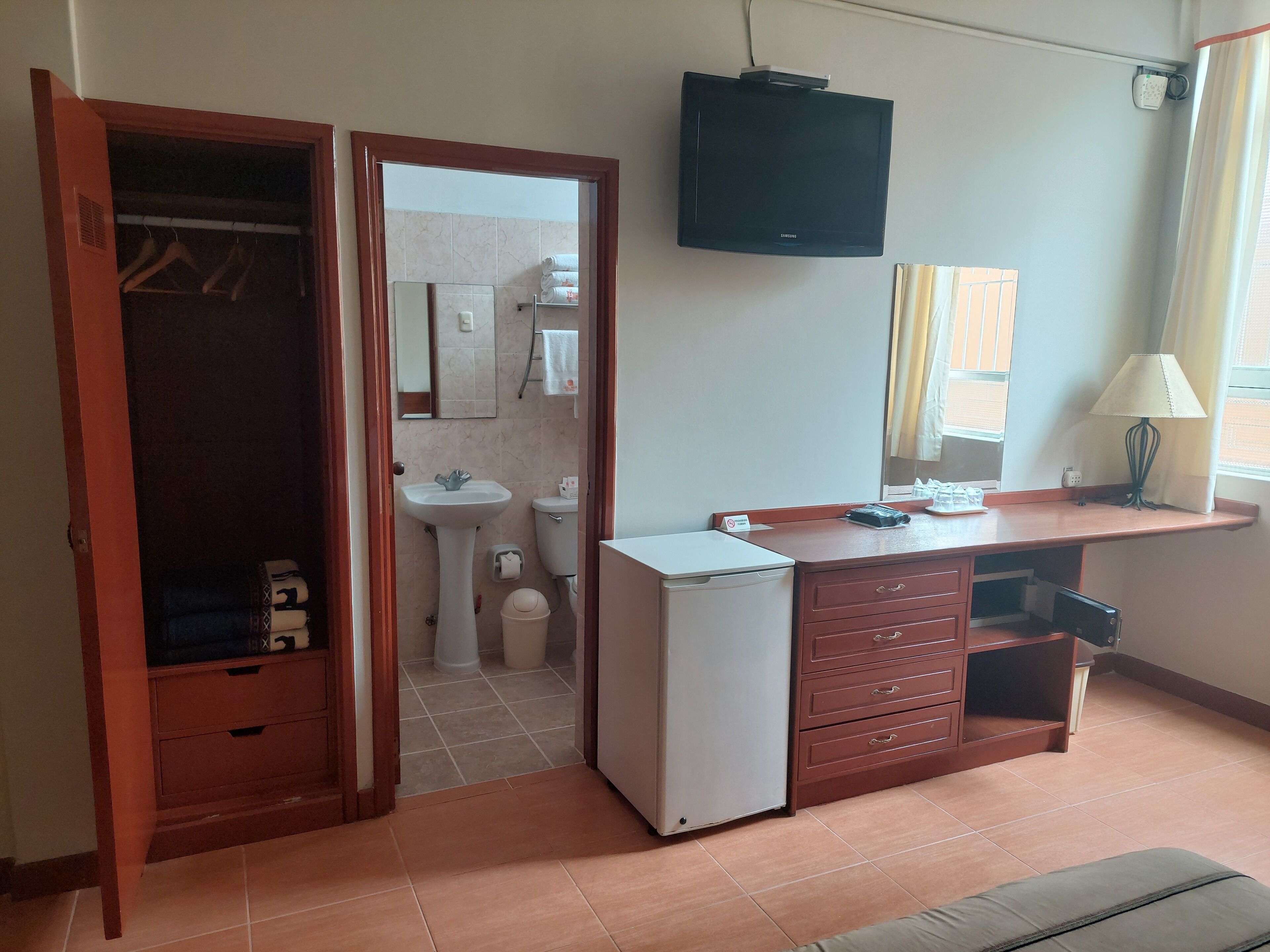 A compact hotel room with a wardrobe, a sink, a toilet, a small refrigerator, a TV, and a bedside table with a lamp.