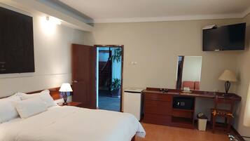 Habitación de hotel con cama, mesita de noche, lámpara, cómoda, espejo, televisor y puerta que da a un pasillo.