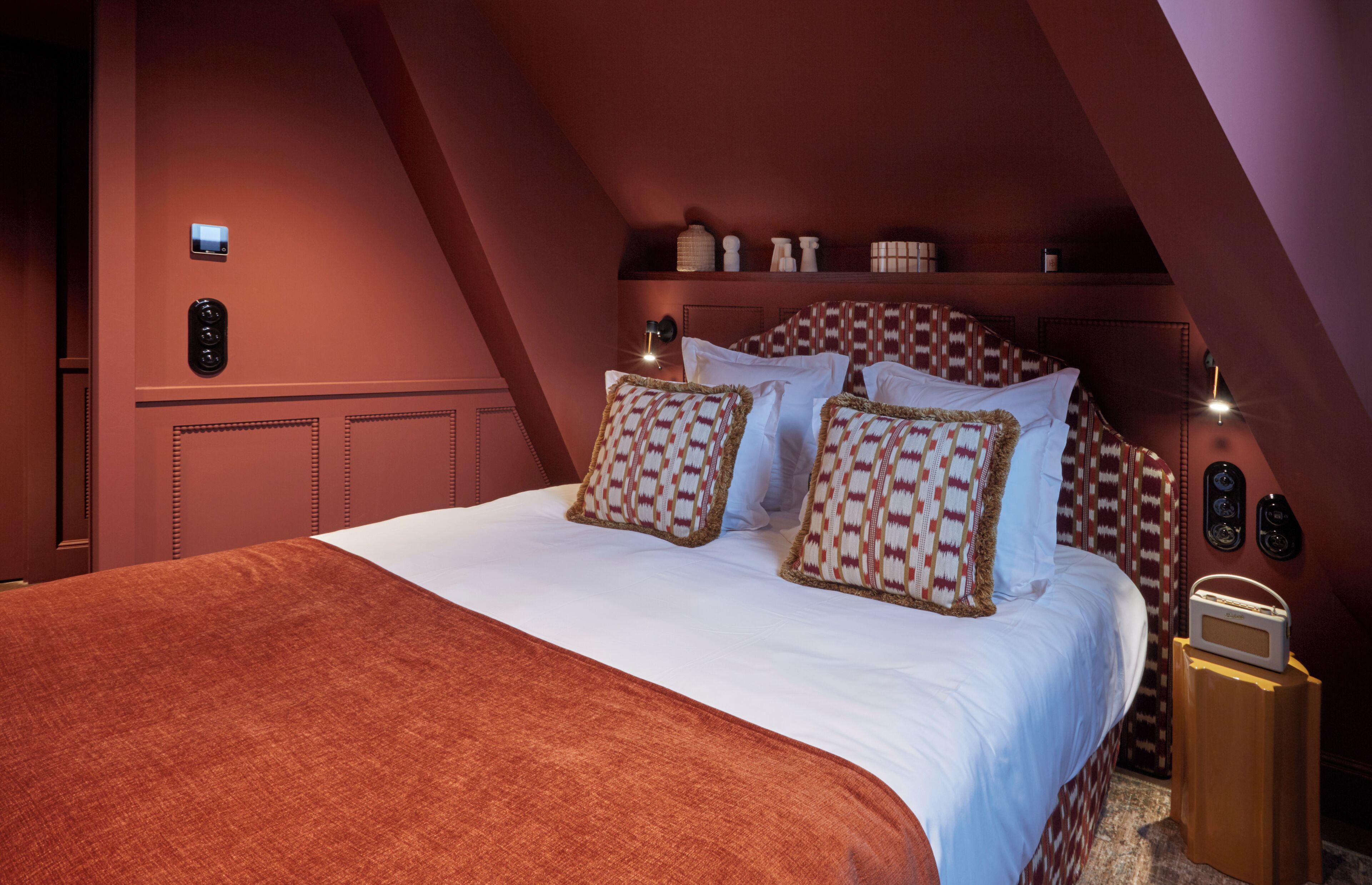 A bedroom with a large bed, patterned pillows, a red accent wall, and a small bedside table with a radio.