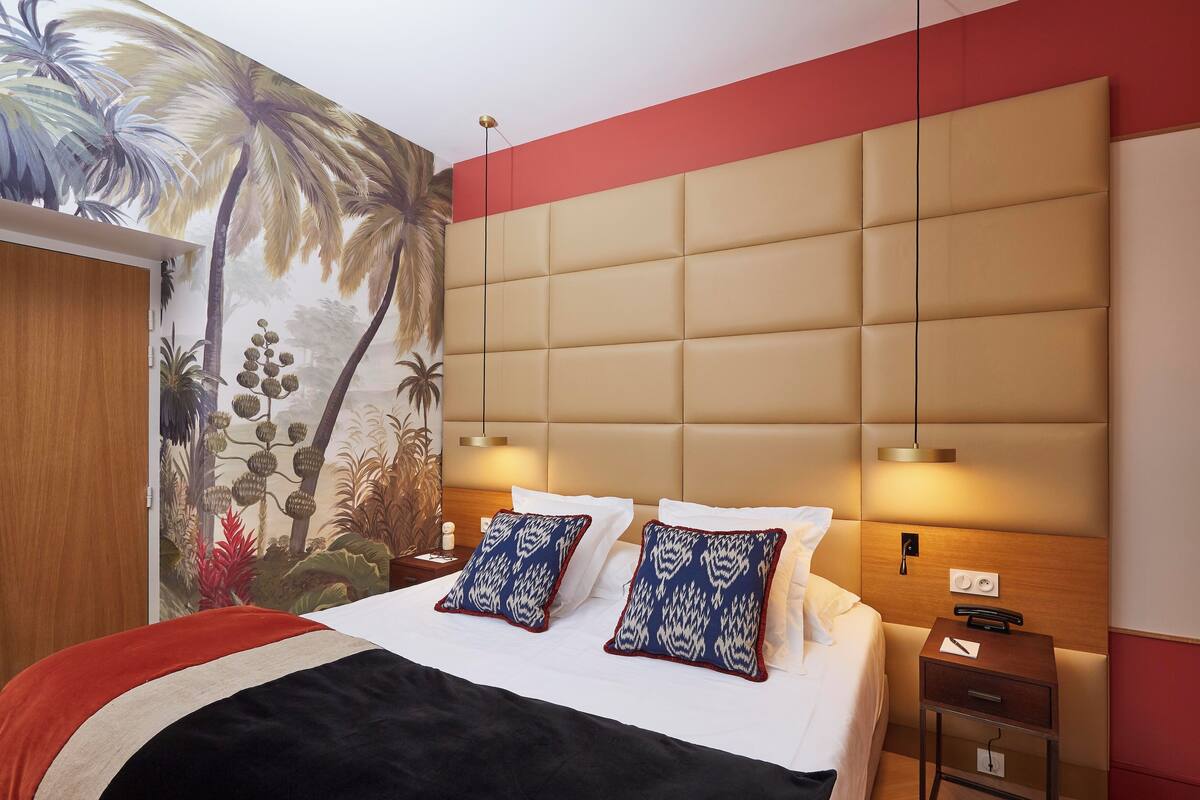 A hotel room with a large bed, a headboard with a light wood finish, two bedside tables with lamps, and a wall mural of tropical plants.