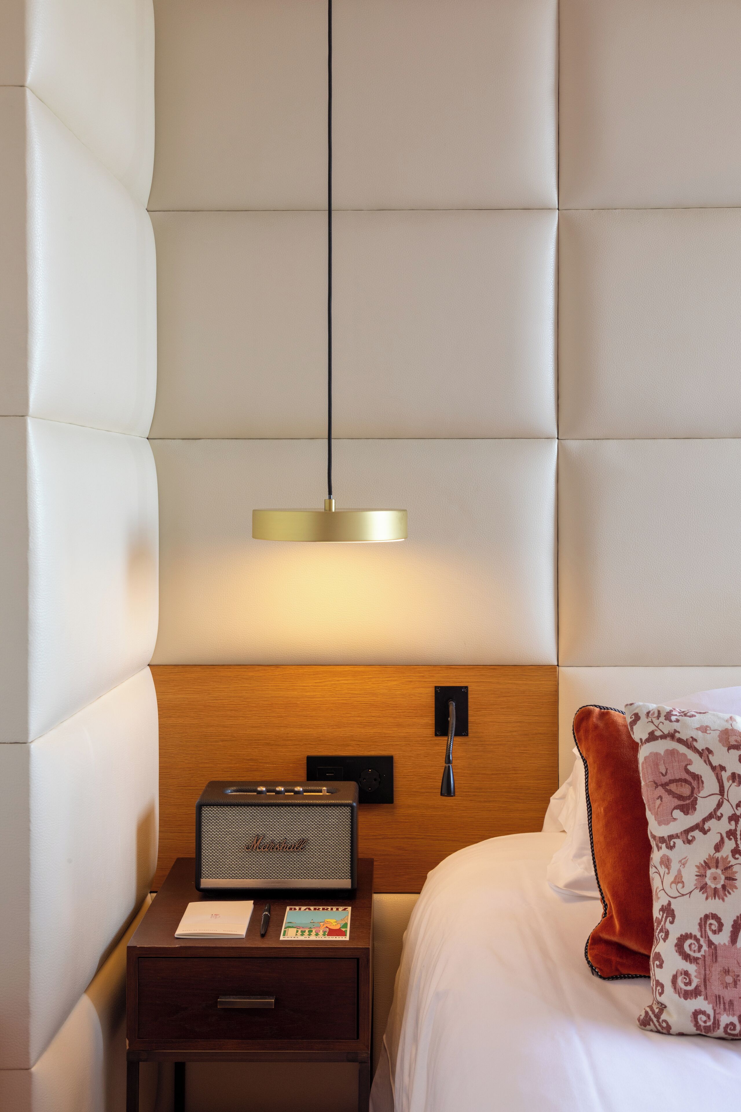 Una cama bien hecha con una almohada estampada, una mesita de noche con una radio, una lámpara y un cajón.