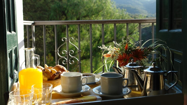 A breakfast setup with a jug of orange juice, cups, a basket of bread, and a vase of flowers on a balcony overlooking a mountainous landscape.