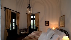 A hotel room with a bed, a desk, and a chair. There is a window with curtains, a hanging light fixture, and a framed picture on the wall.