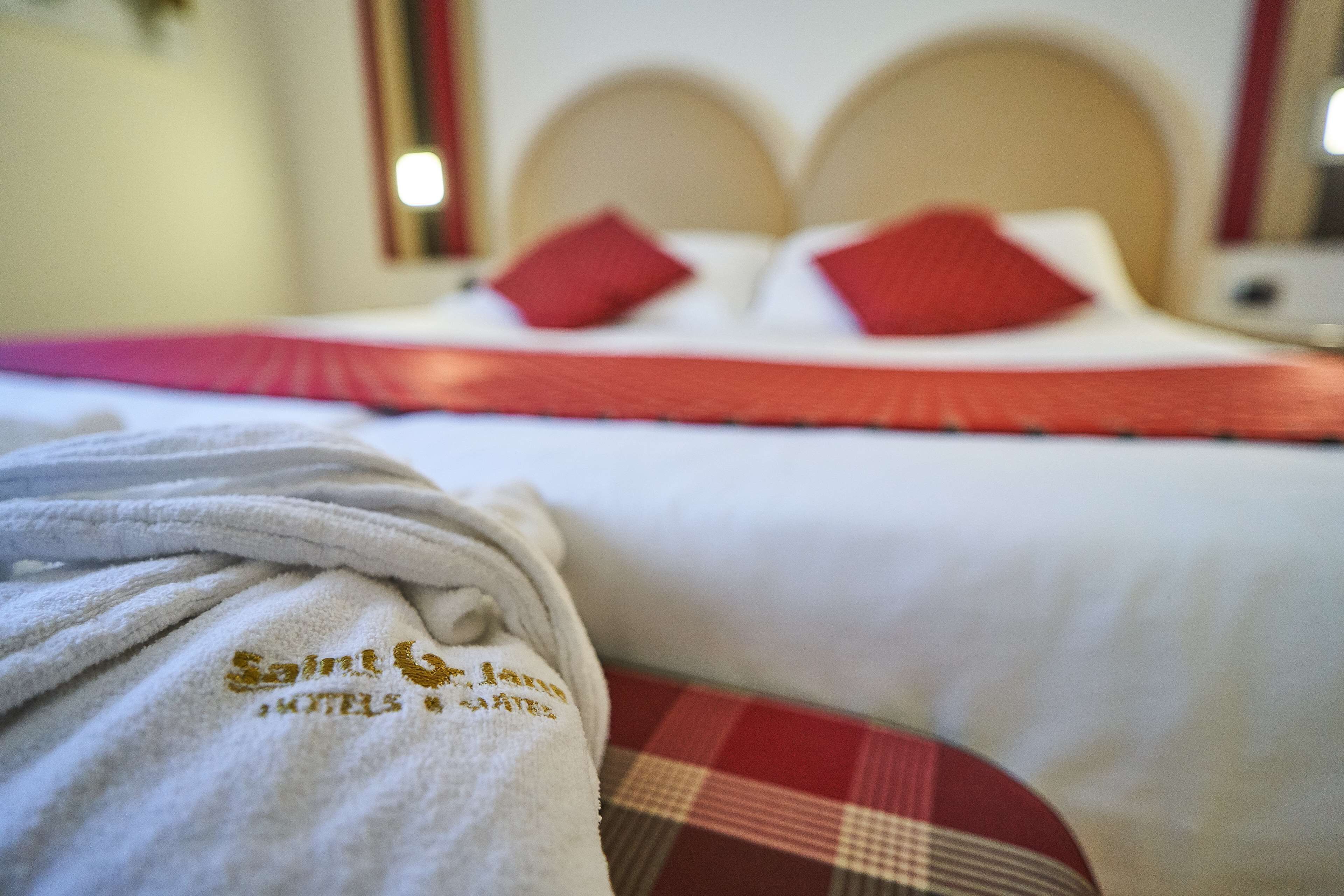 A hotel room with a bed featuring a red and white checkered bedspread, a white headboard, and red pillows. A white bathrobe with embroidered text is draped over the bed.