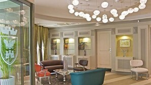Una sala de estar moderna con un sofá azul, un sillón marrón y un sillón verde azulado. Se observa una partición de vidrio con un diseño de escudo de armas. Un candelabro con múltiples luces esféricas cuelga del techo.