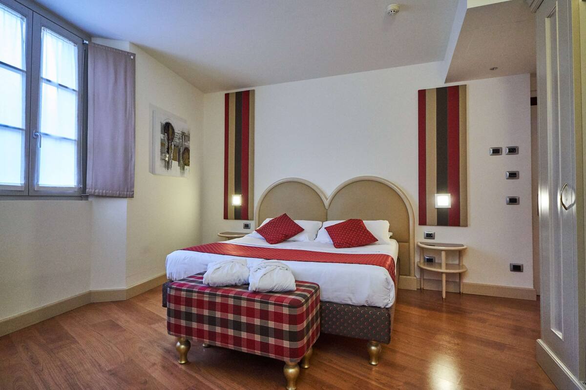 A hotel room with a large bed, two bedside tables, a small stool, and a window with curtains.