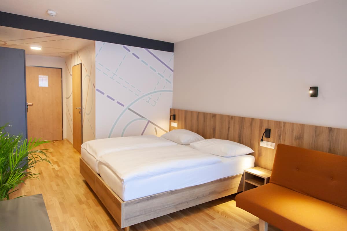 Una habitación de hotel moderna con suelo de madera, una cama grande con ropa blanca, un cabecero de madera, una mesita de noche con lámpara y una pequeña zona de estar con un cojín.