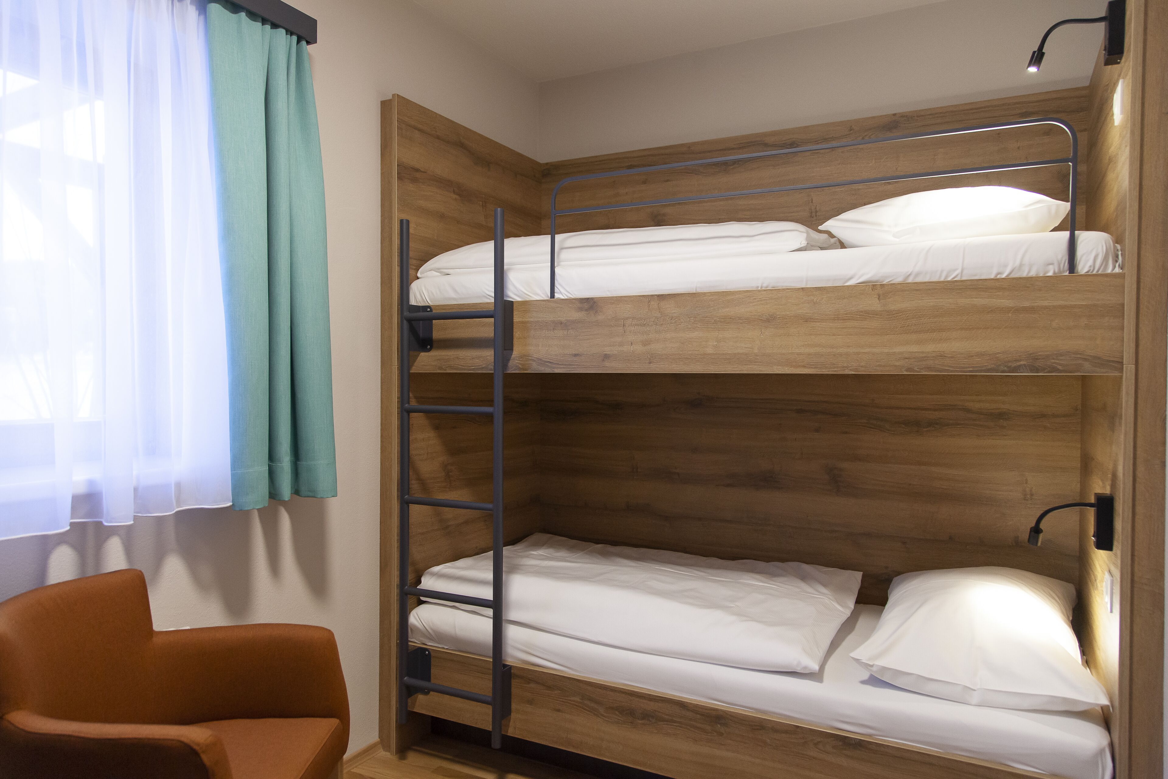 A bunk bed room with a ladder, a chair, and a window with curtains.