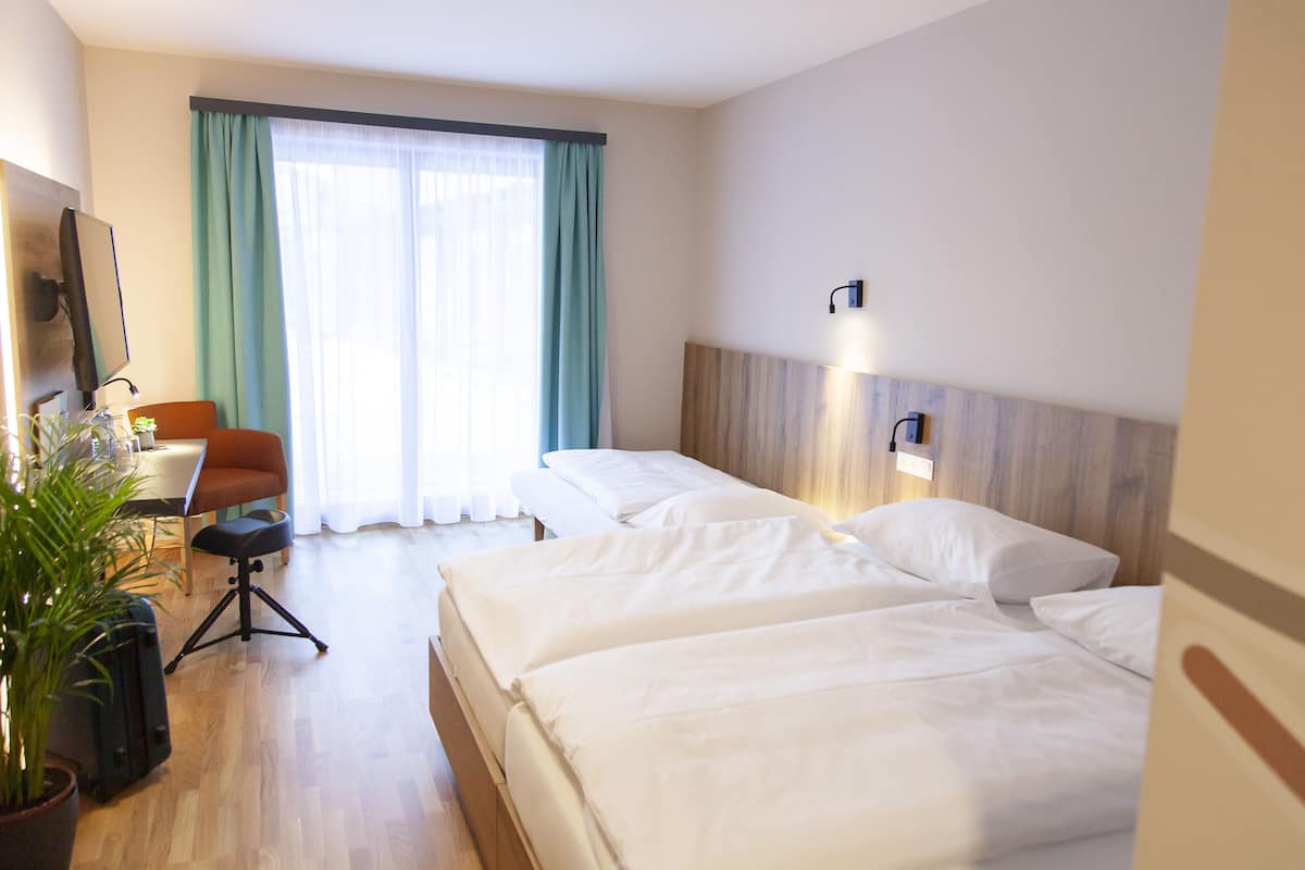 Habitación de hotel con dos camas, un escritorio con silla, una planta en maceta y un ventanal con cortinas.