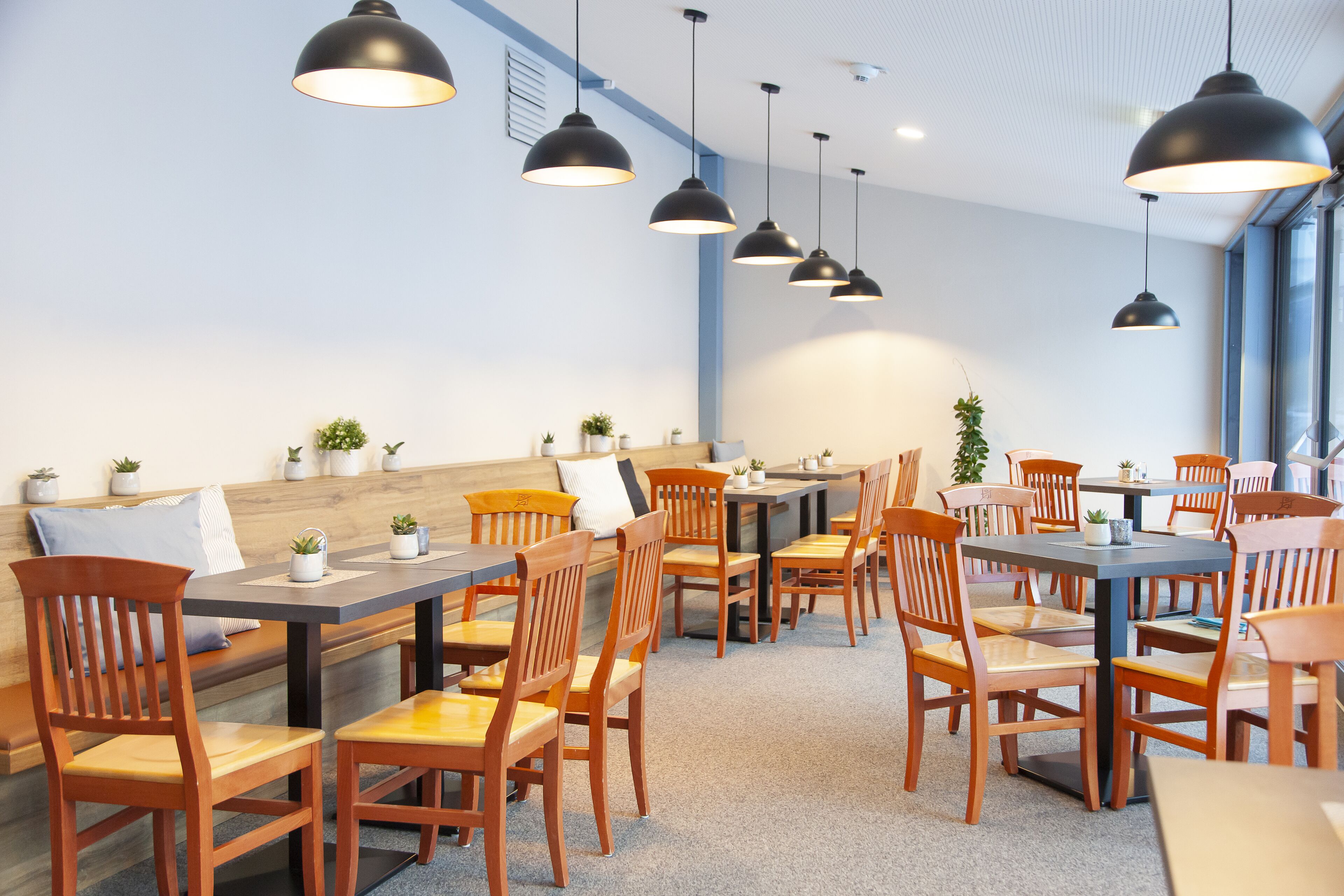 A modern dining area with wooden tables and chairs, black pendant lights, and small potted plants.