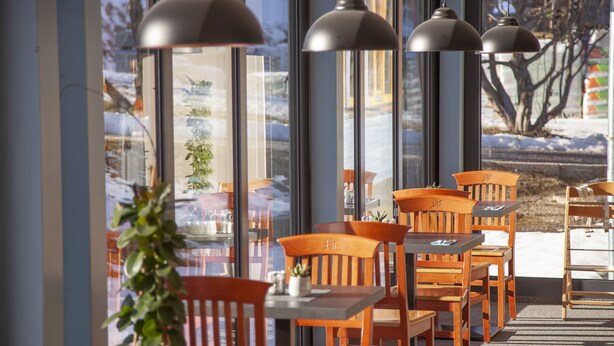 An outdoor seating area with wooden tables and chairs, large windows, and hanging pendant lights. - JUFA Hotel Erlaufsee (Mariazell)