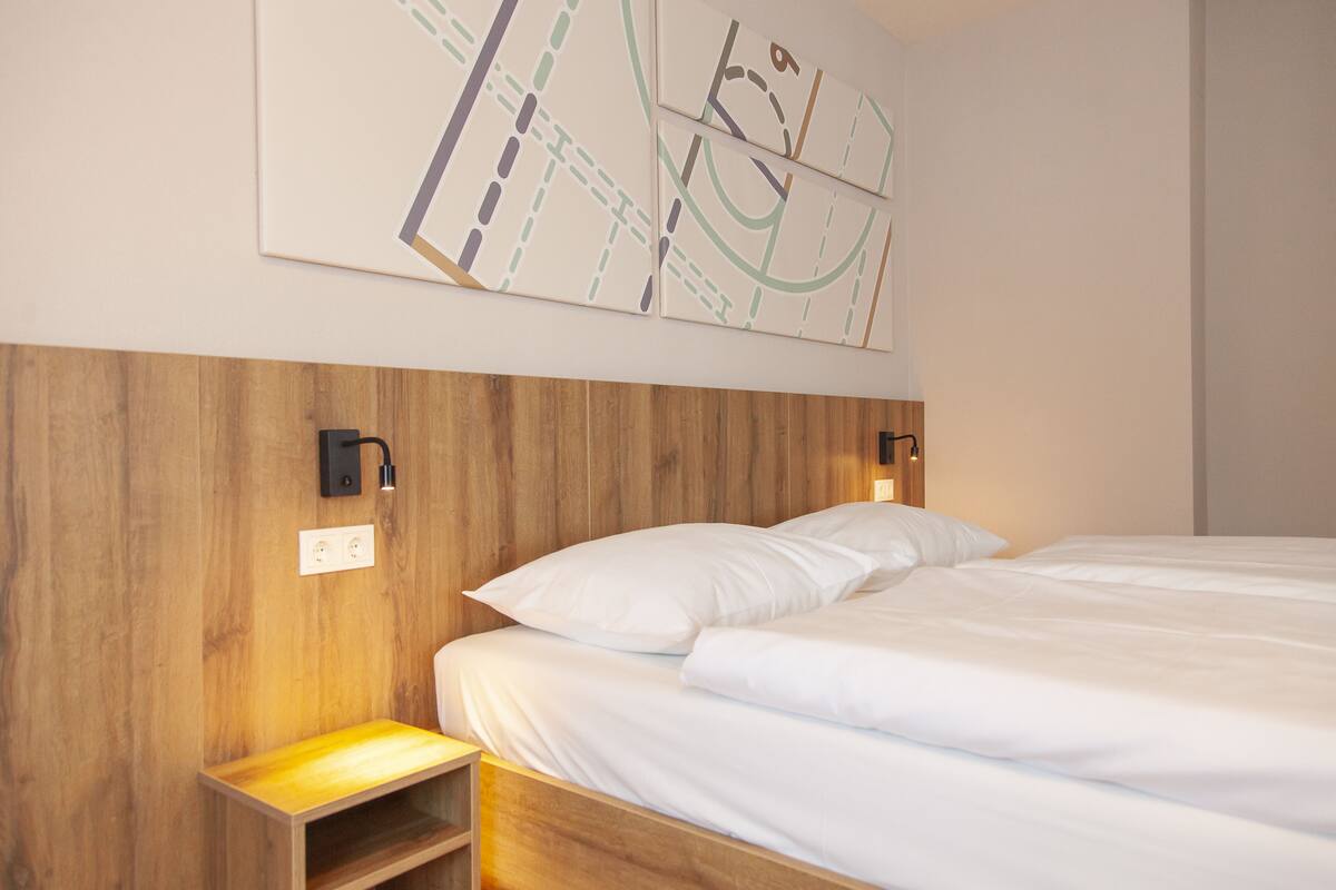 Una cama bien tendida con sábanas blancas, cabecera de madera y lámpara de pared.