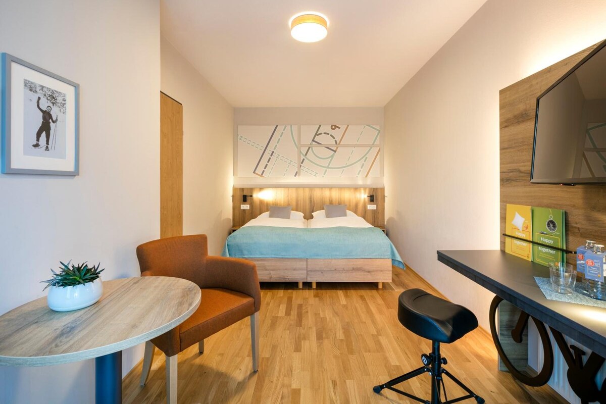 A modern hotel room with a bed, a desk, a chair, a small table, and a potted plant.