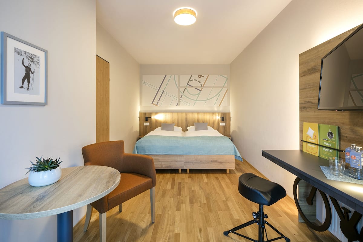 Una habitación de hotel moderna con piso de madera, una cama con cabecera azul, un escritorio con silla, una mesita con una planta y un cuadro en la pared.