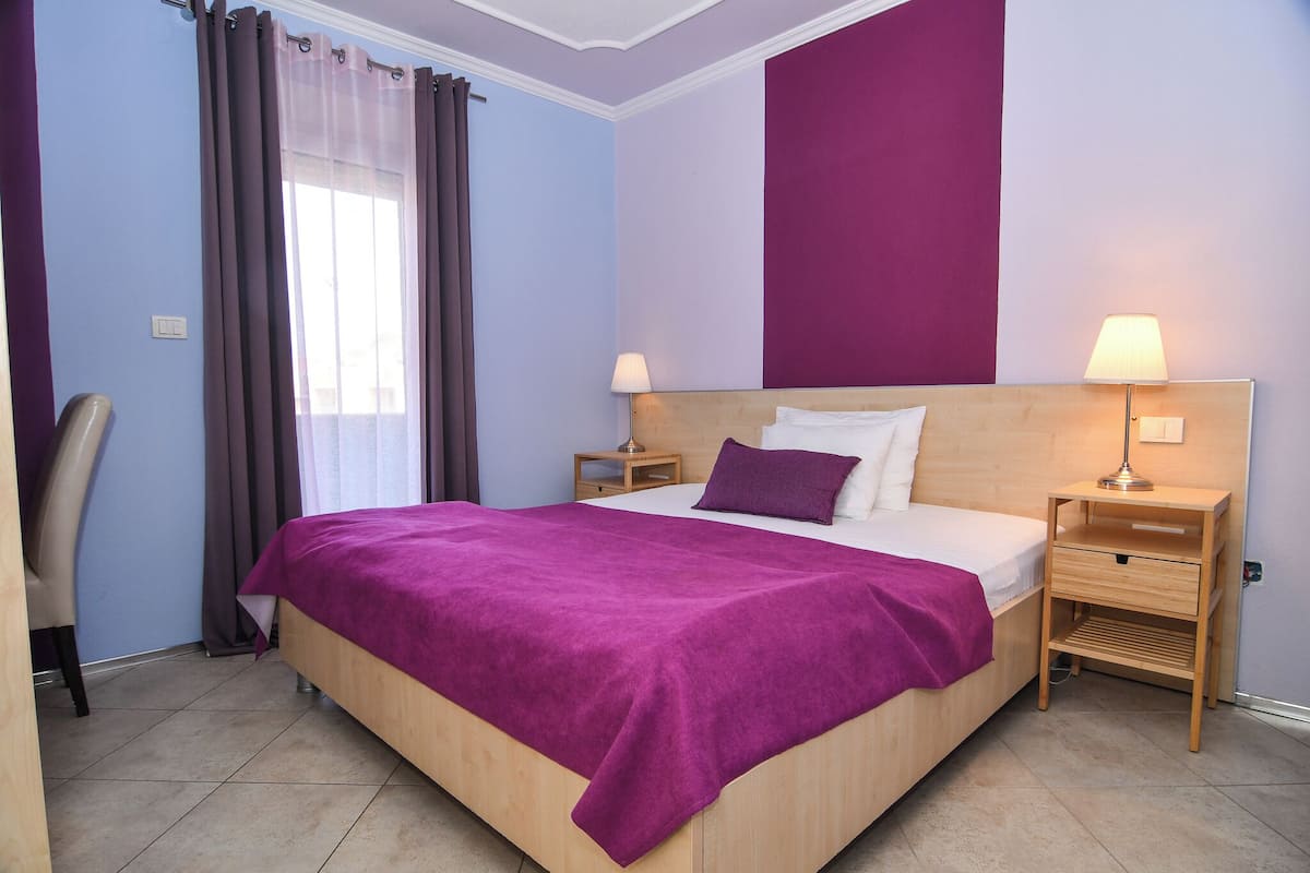A bedroom with a bed, two bedside tables, a chair, a window with curtains, and a wall with a purple panel.