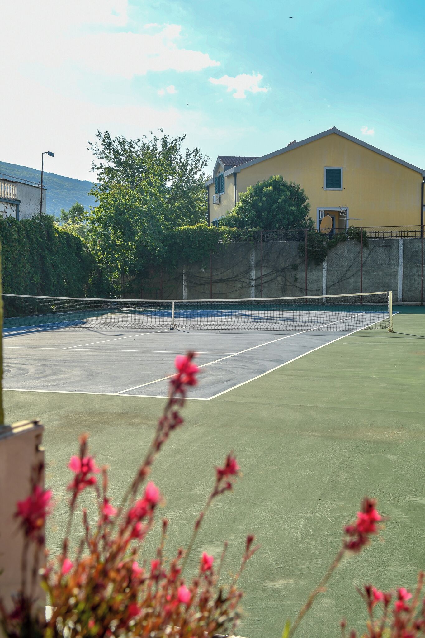 An outdoor tennis court with a net, surrounded by greenery and a building in the background.