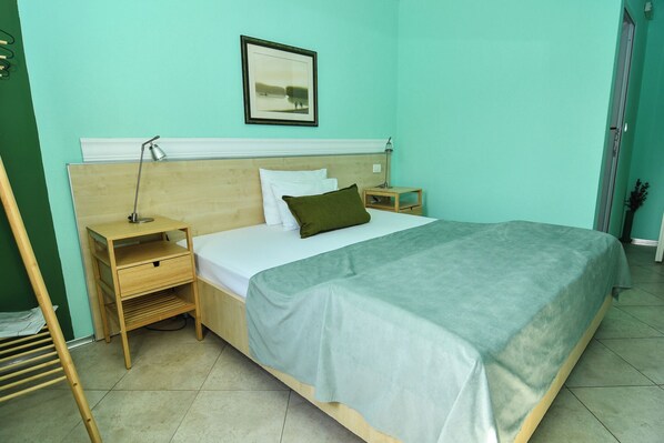 A neatly made bed with a green bedspread, a wooden nightstand with a lamp, a framed picture on the wall, and a wooden floor. - Hotel Aruba (Lastva Grbaljska)