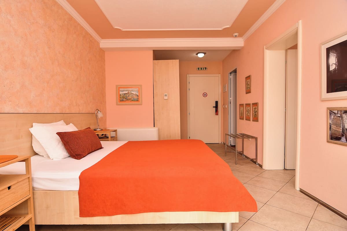 A hotel room with a bed, bedside tables, a nightstand, a door with an exit sign, and a hallway with a mirror and framed pictures.