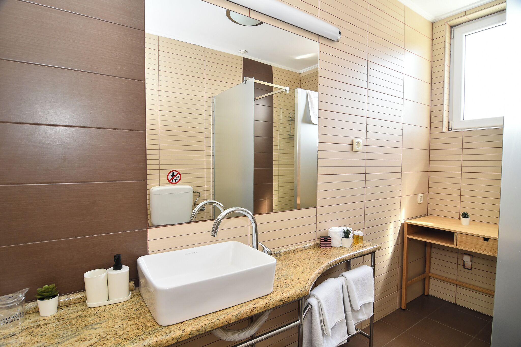 Un baño moderno con lavabo, espejo, toallero y un pequeño escritorio con una planta.
