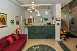 Zona de recepción de un hotel con un sofá rojo, un mostrador de recepción y una escalera que sube a otro piso.