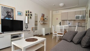 Una sala de estar moderna con televisor de pantalla plana, muebles blancos, un comedor con sillas y una cocina con armarios.