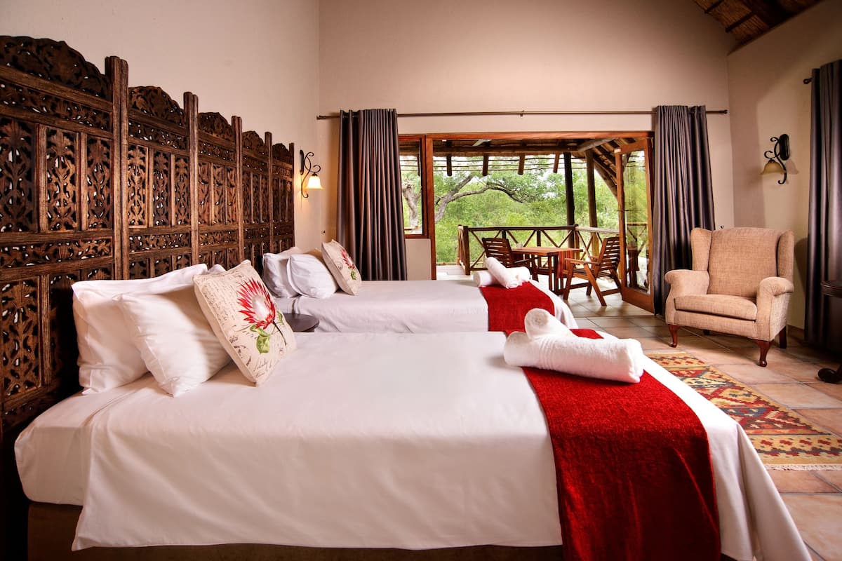 A hotel room with two beds, a wooden headboard, a red blanket, a chair, and a view of the outdoors.