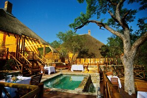 A rustic outdoor dining area with wooden furniture, a pool, and thatched-roof buildings. - La Kruger Lifestyle Lodge (Nkomazi)