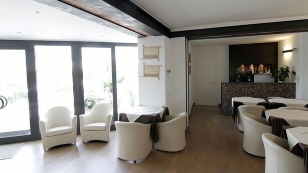 A modern dining area with white chairs and tables, a kitchen in the background, and large windows allowing natural light.