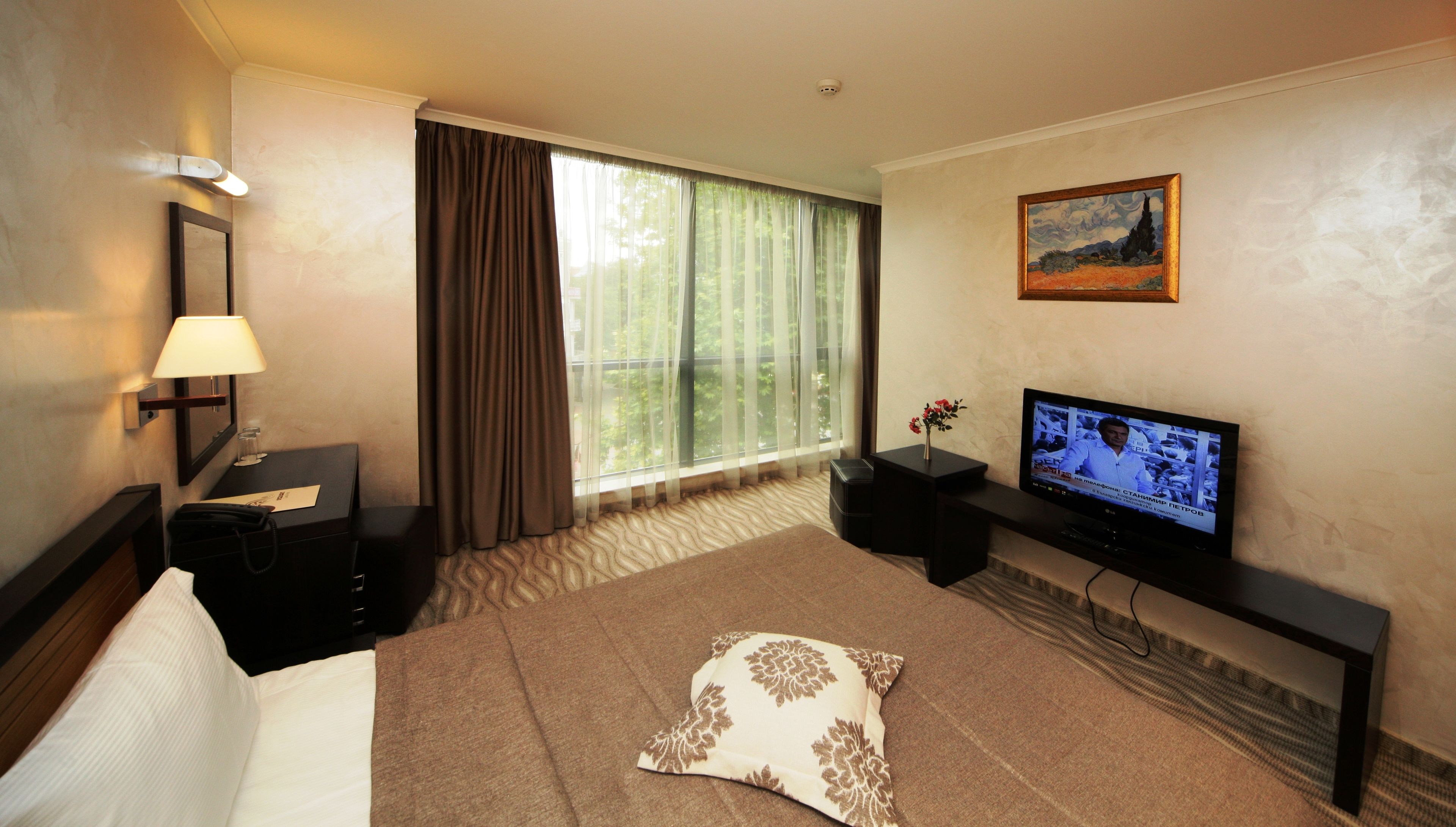 Quarto de hotel com cama, escrivaninha com luminária, televisão e um quadro na parede.