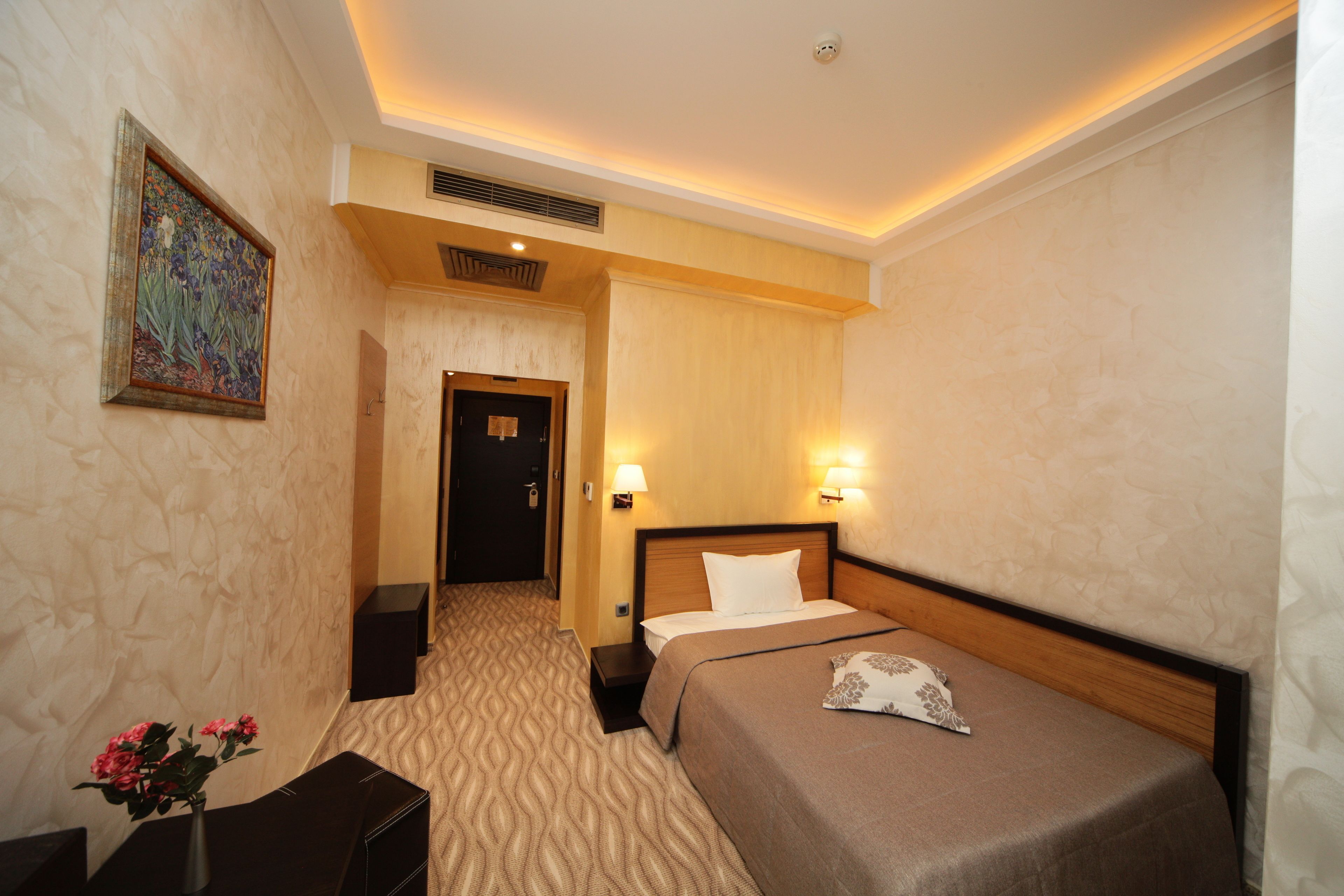 A hotel room with a bed, bedside lamps, a nightstand, a vase with flowers, a painting on the wall, and a door leading to another room.