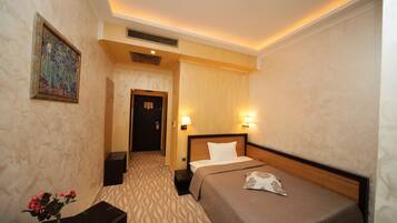 A hotel room with a bed, bedside lamps, a nightstand, a vase with flowers, a painting on the wall, and a door leading to another room.