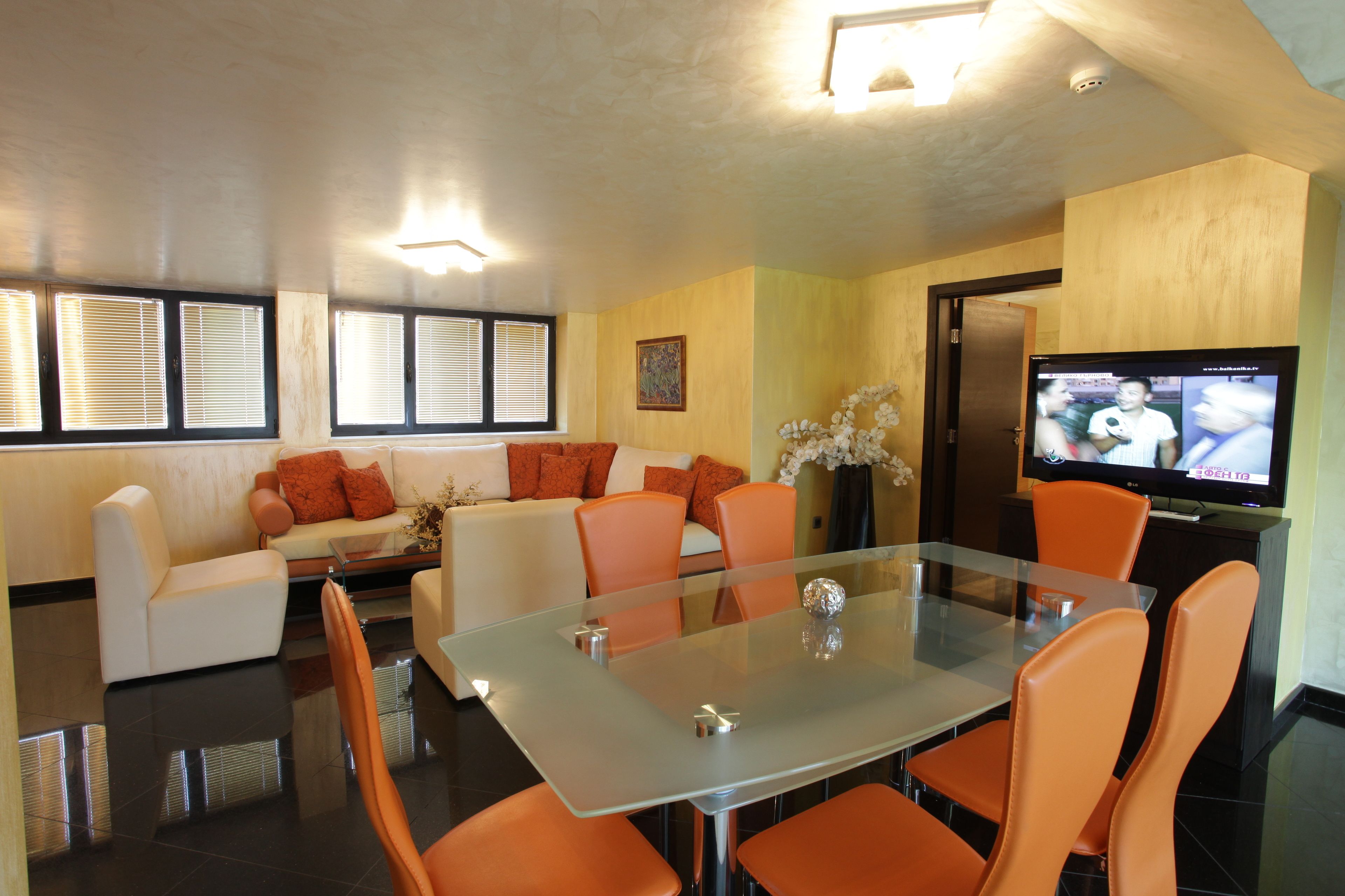 Una sala moderna con una mesa de comedor de cristal, sillas naranjas, un sofá blanco y un televisor sobre un mueble.