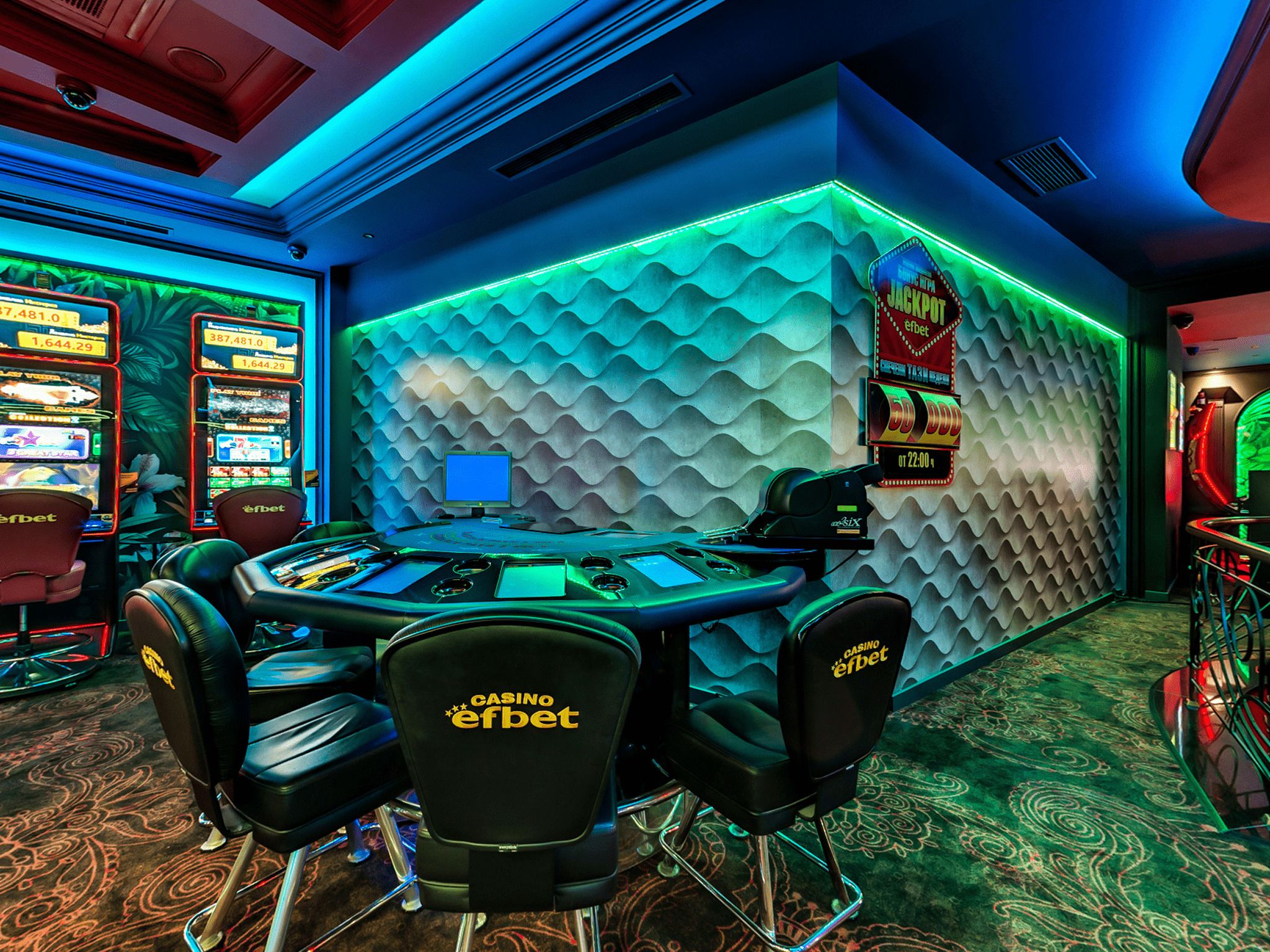 Un espace de jeux de casino comprenant plusieurs machines à sous, une table centrale entourée de chaises et un mur à la texture particulière.
