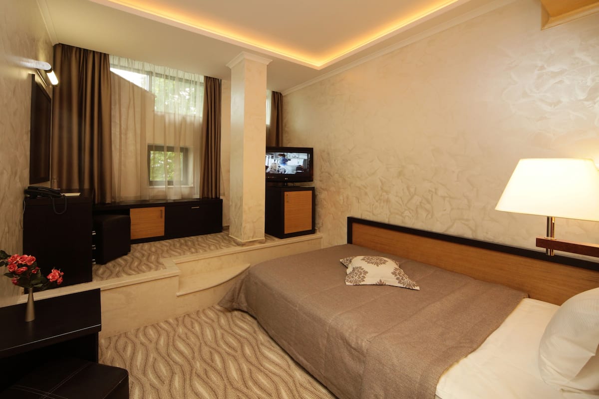 A hotel room with a bed, a TV, a nightstand, a lamp, a window with curtains, and a painting on the wall.