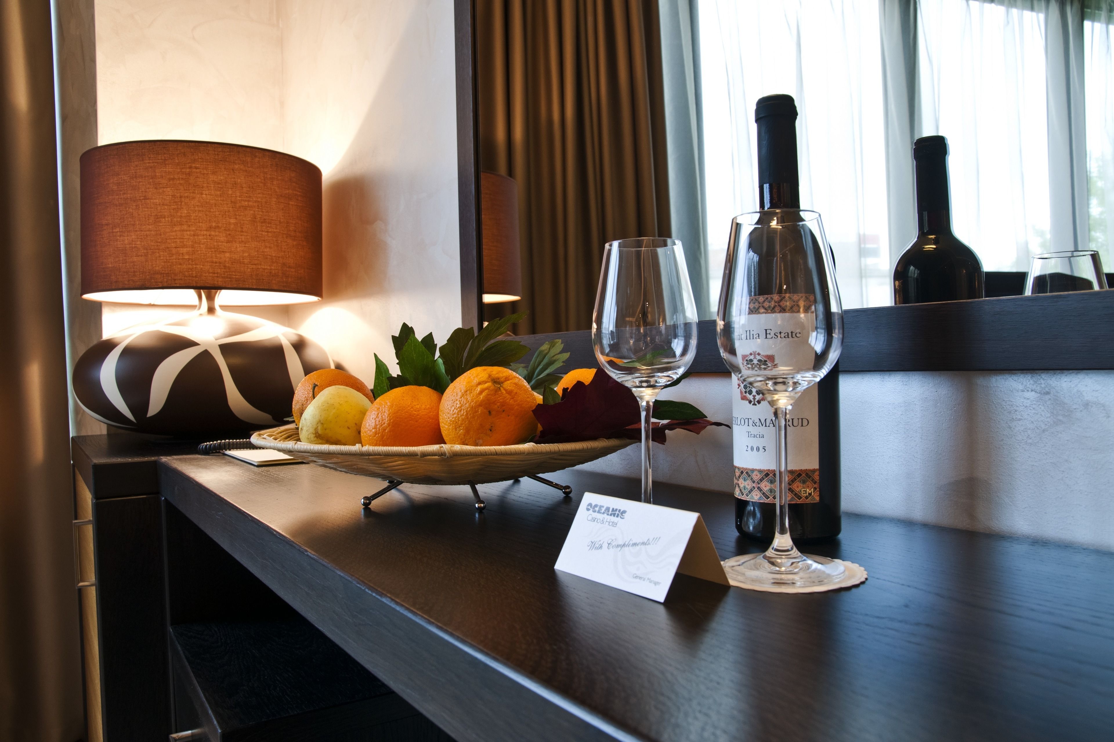 Ein Hotelzimmer mit Schreibtisch aus Holz, einer Lampe, einer Obstschale mit Orangen, einer Weinflasche und einem Weinglas.