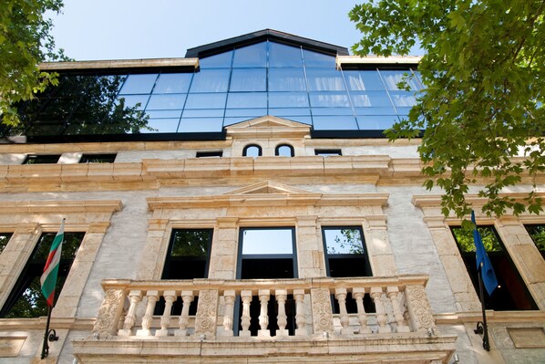 A building with classical architecture, featuring a prominent balcony with ornate balustrades and a large glass facade reflecting the sky.