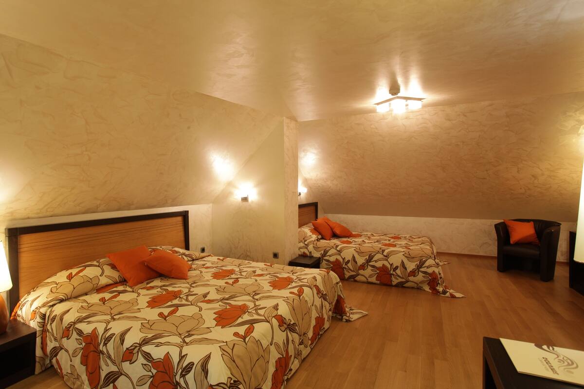 A hotel room with two beds, each with floral bedding and orange pillows. There are bedside tables with lamps, a wooden floor, and a chair in the corner.